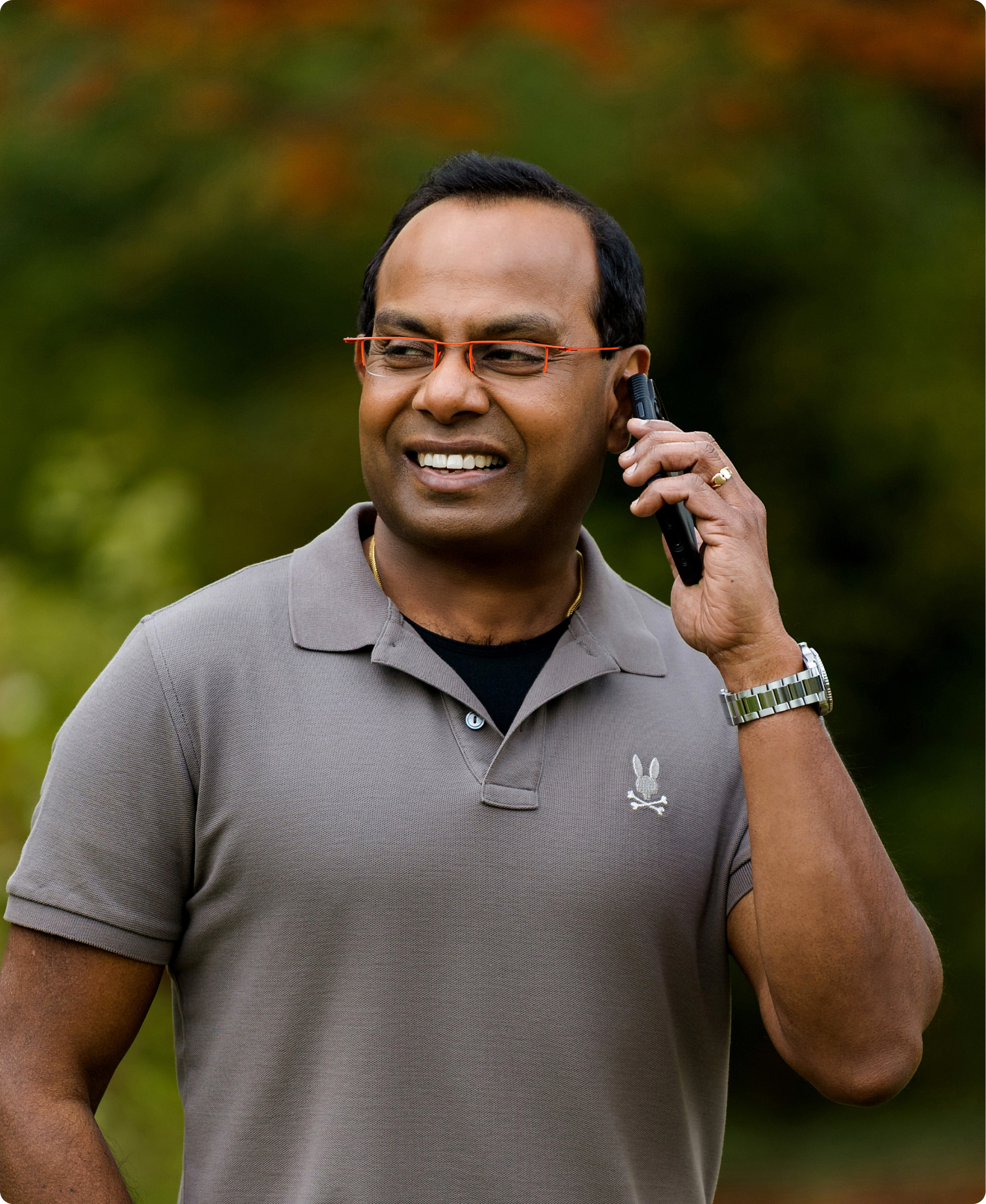 Smiling man outdoors wearing orange-framed glasses, speaking into a handheld headset device while wearing a gray polo shirt and watch.