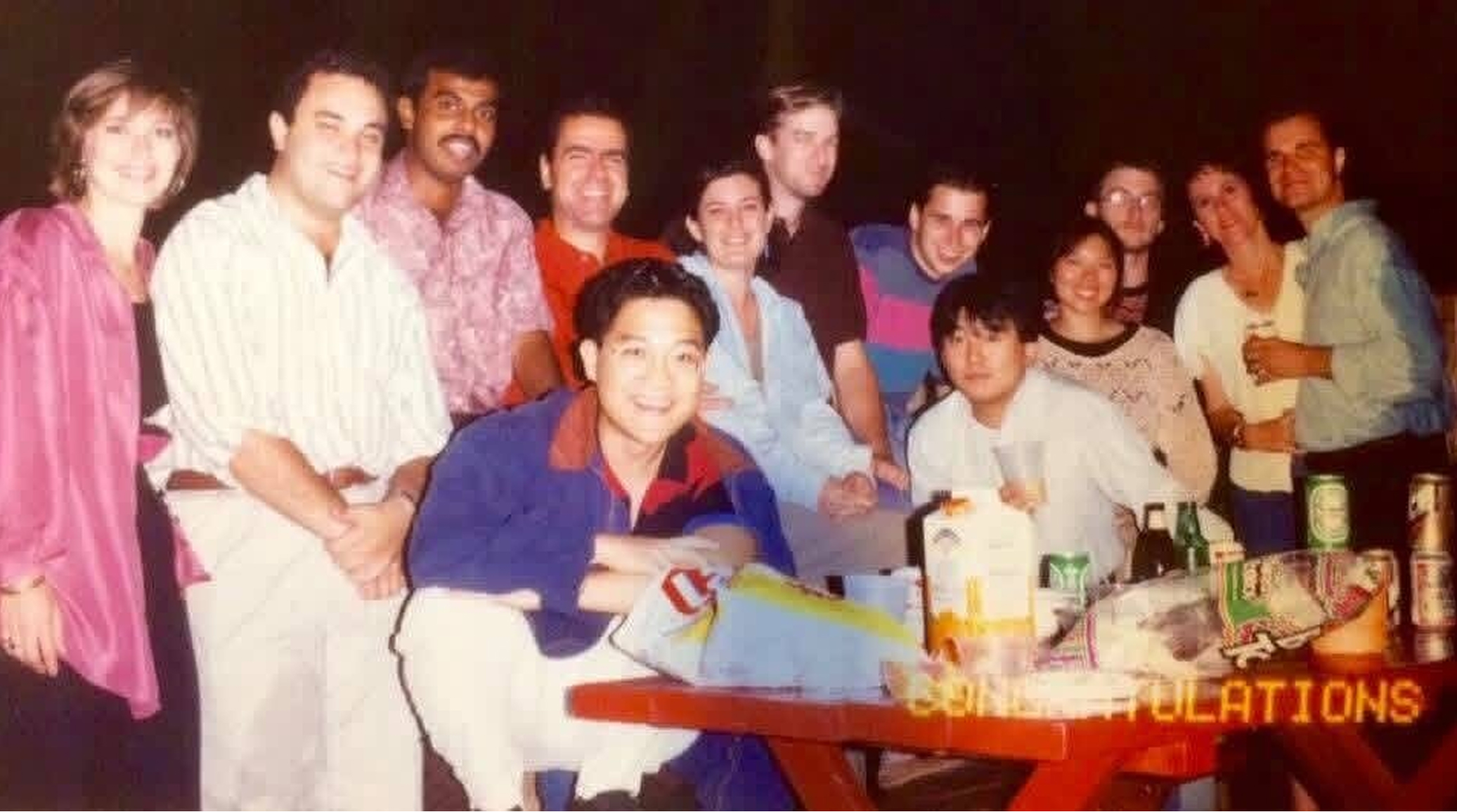 Group of people smiling and posing for a photo at a party, with a celebratory banner reading 'CONGRATULATIONS' in the foreground and drinks on the table.