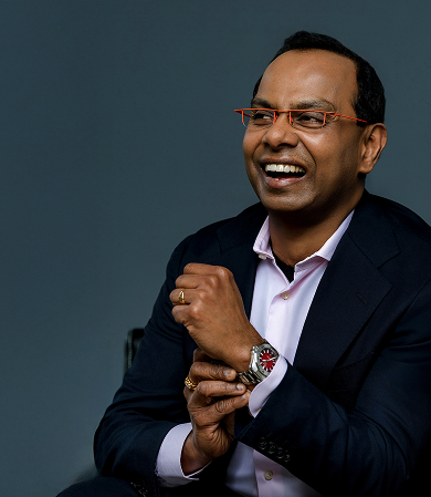 Portrait of a man in a navy suit and pink shirt, smiling with hands clasped, wearing an orange wristwatch and orange glasses.